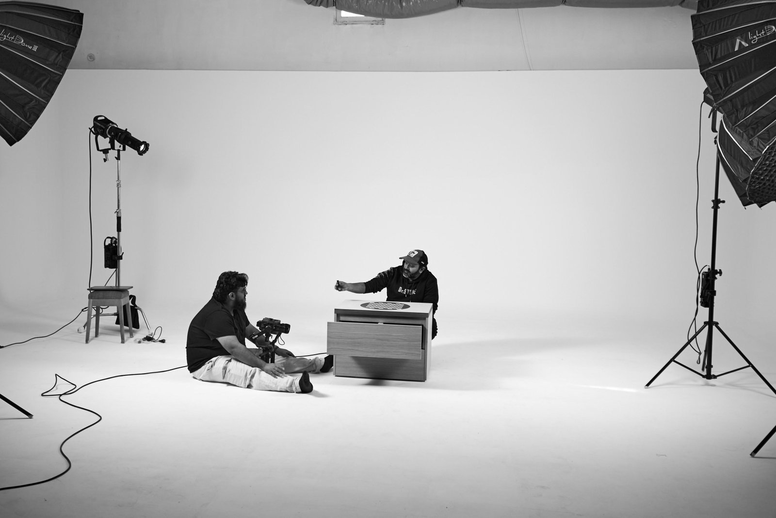 Équipe en préparation dans un studio cyclorama : un opérateur assis au sol tient une caméra sur stabilisateur, face à un intervenant qui ajuste un élément de décor posé sur une commode. Deux boîtes à lumière et un projecteur encadrent le plateau.