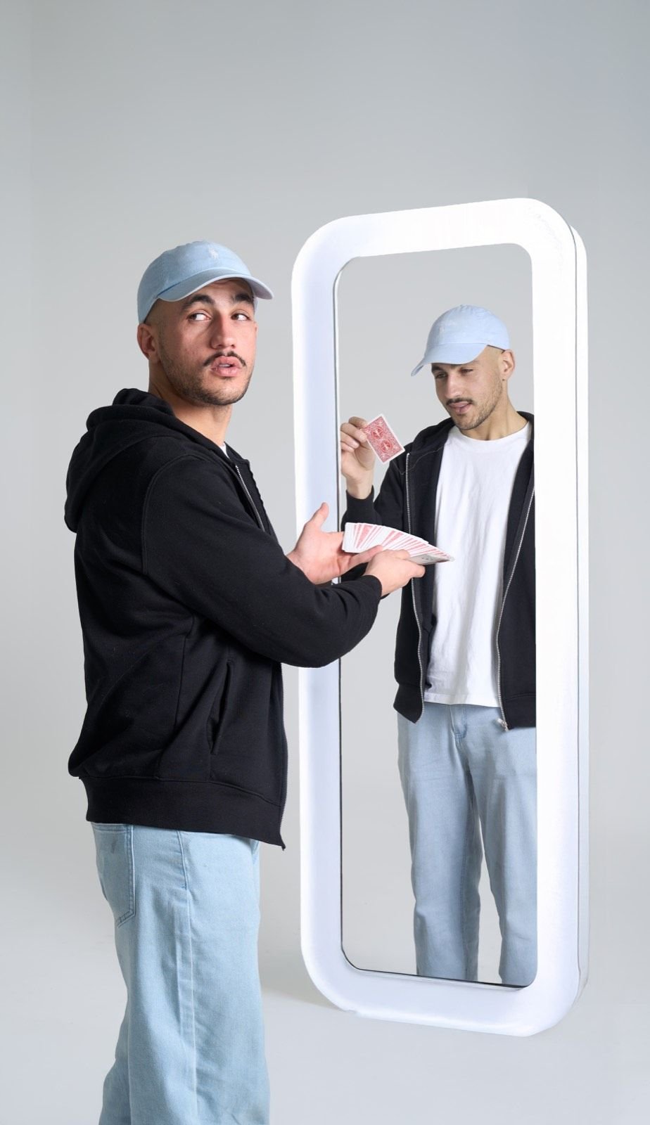 Un magicien en casquette présente un éventail de cartes face à un grand miroir sur pied ; son reflet, décalé, tient une carte isolée. Fond de studio neutre.